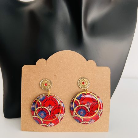 Boucles d'oreilles rondes en tissu wax rouge et bleu avec métal doré, affichées sur un présentoir en carton.